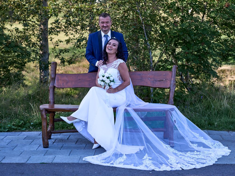 A couple poses on a wooden bench outdoors in wedding attire with a flowing white dress and blue suit.