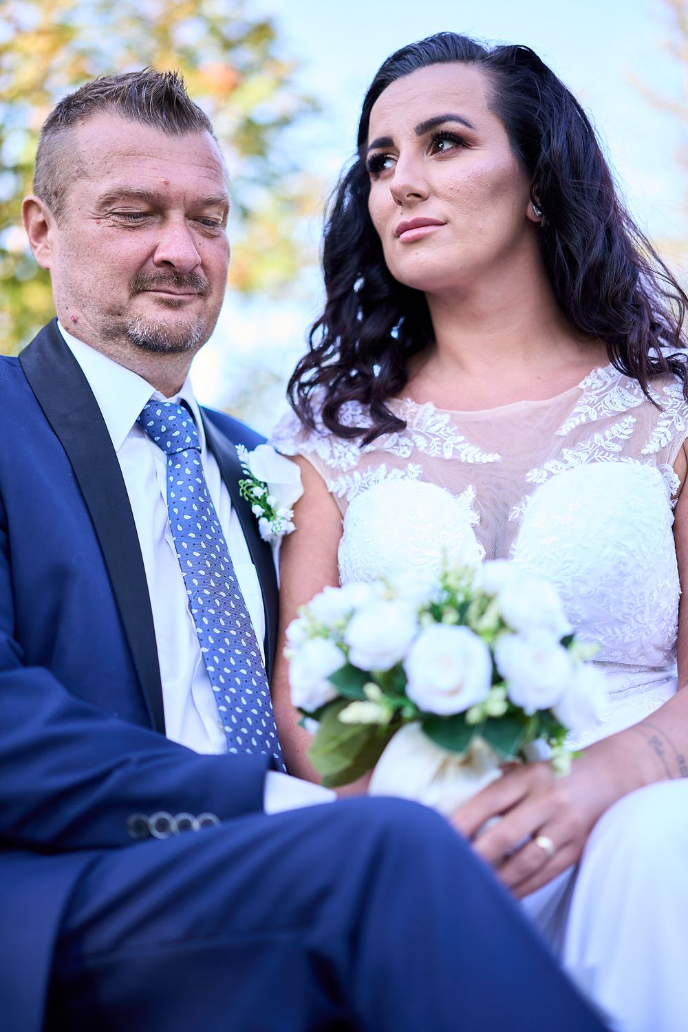A newlywed couple poses outdoors with the bride in white lace holding a bouquet and groom in navy suit with blue tie.