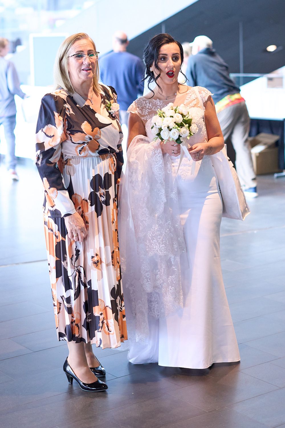 Two people stand together at a formal event, one wearing a floral dress and one in a white wedding gown holding a bouquet.