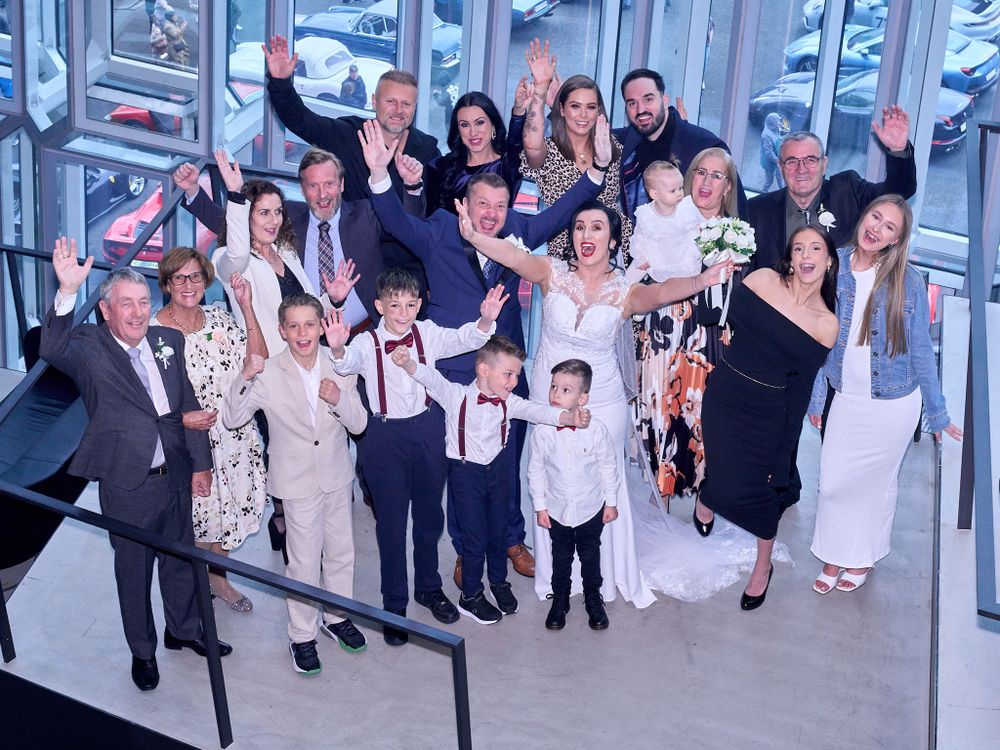 A festive group photo of wedding guests and a bride in white dress gathered together in a modern building interior.