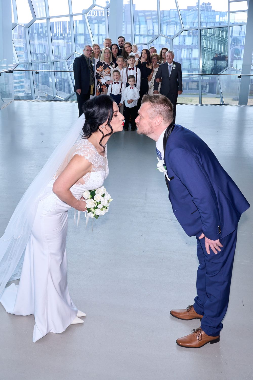 A bride and groom lean in for a playful kiss while wedding guests watch from behind in a modern indoor venue.