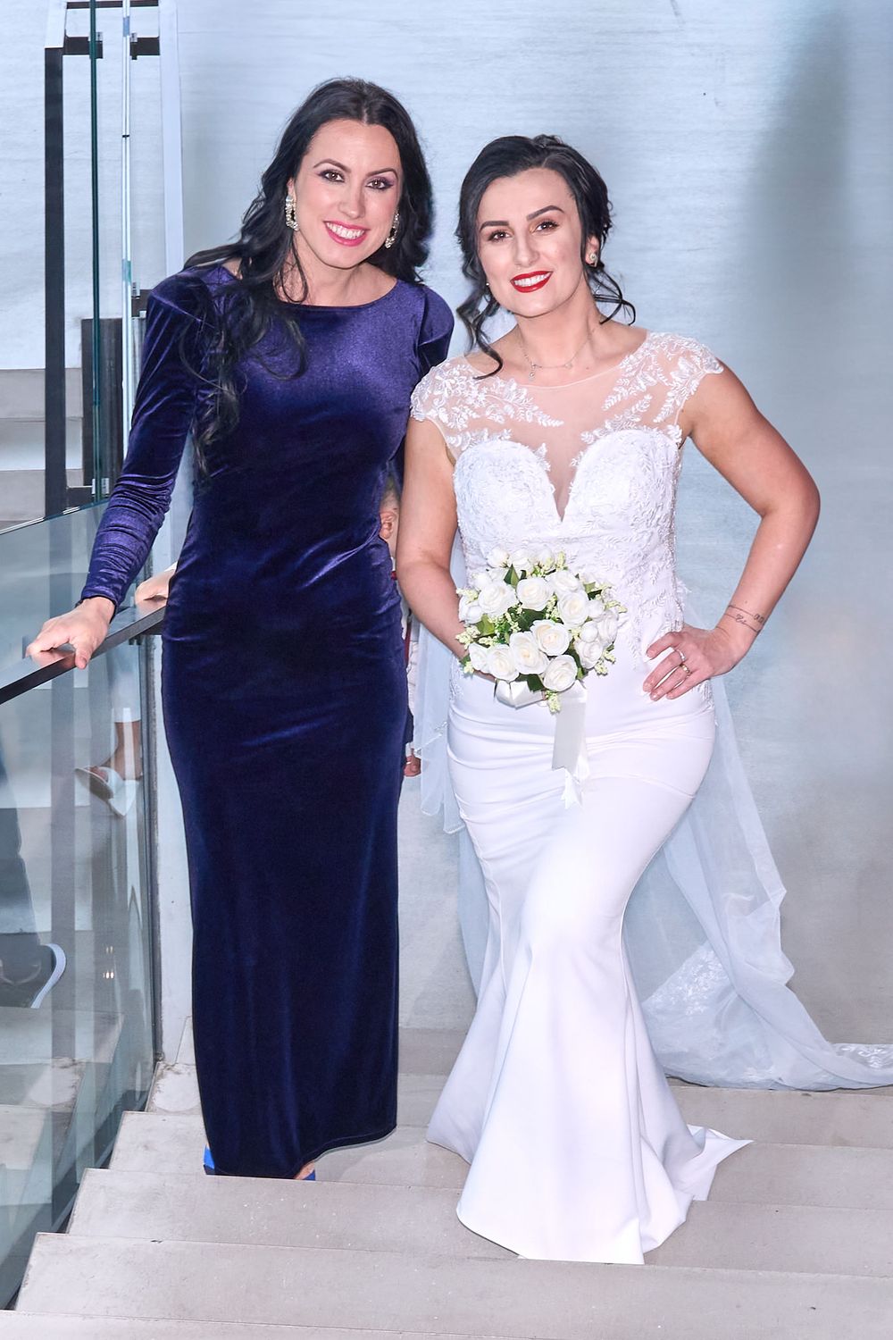 Two women pose together at a formal event, one wearing a navy velvet gown and one in a white lace wedding dress with bouquet.