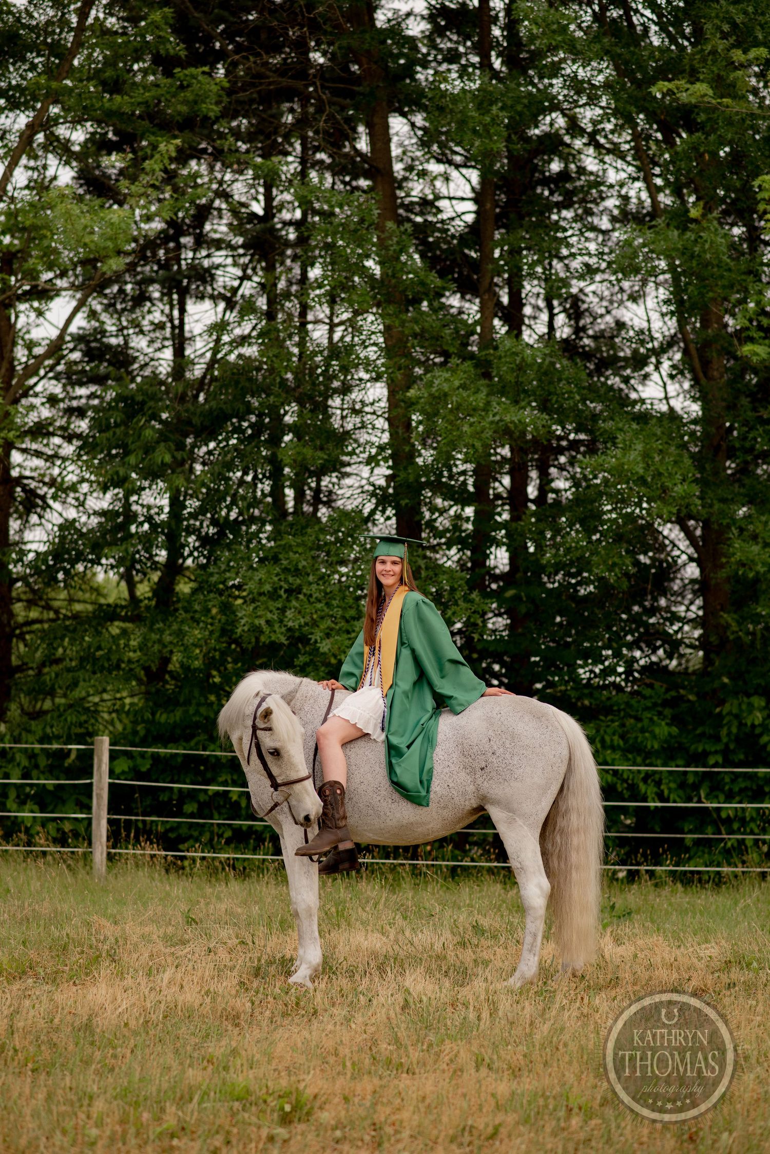 Graduate in green robes sits mounted on a white horse in fenced pasture.