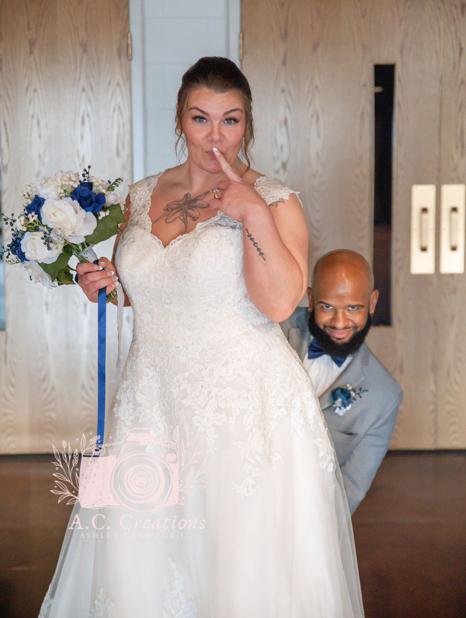 A bride in a white lace dress holds a blue and white bouquet while posing playfully with her partner.