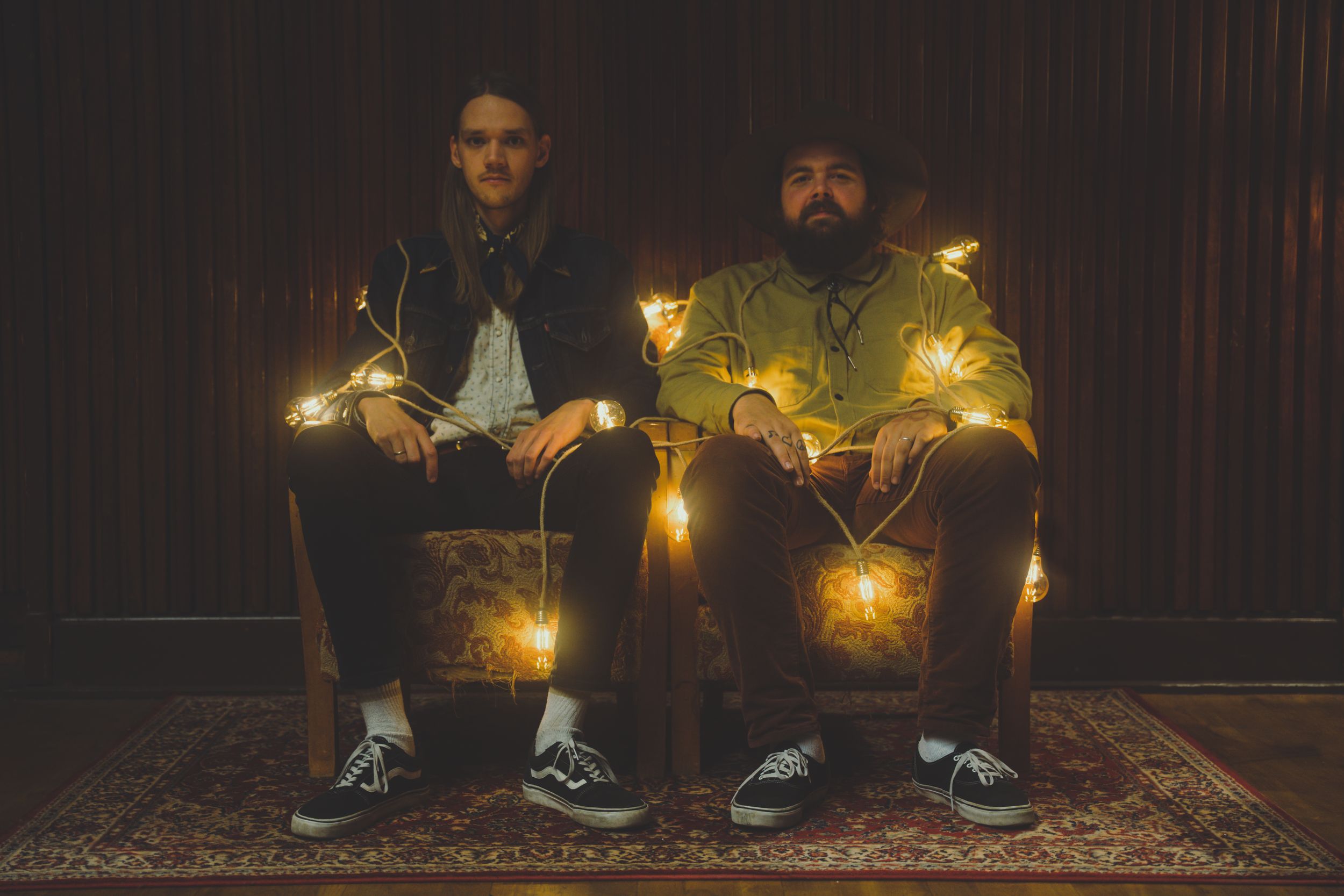 Two figures sitting on chairs surrounded by glowing string lights in a dark room.