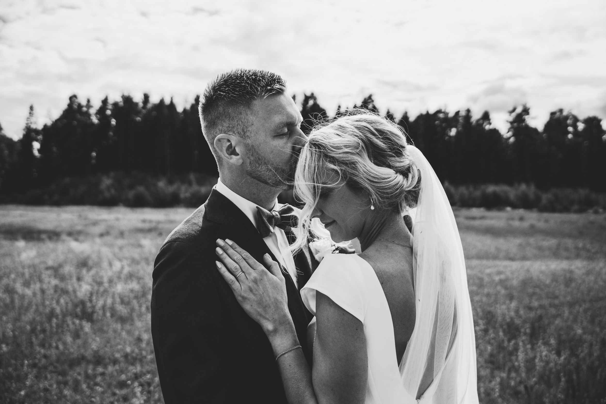 Black and white wedding photo of bride and groom sharing an intimate moment in a field with trees in background.