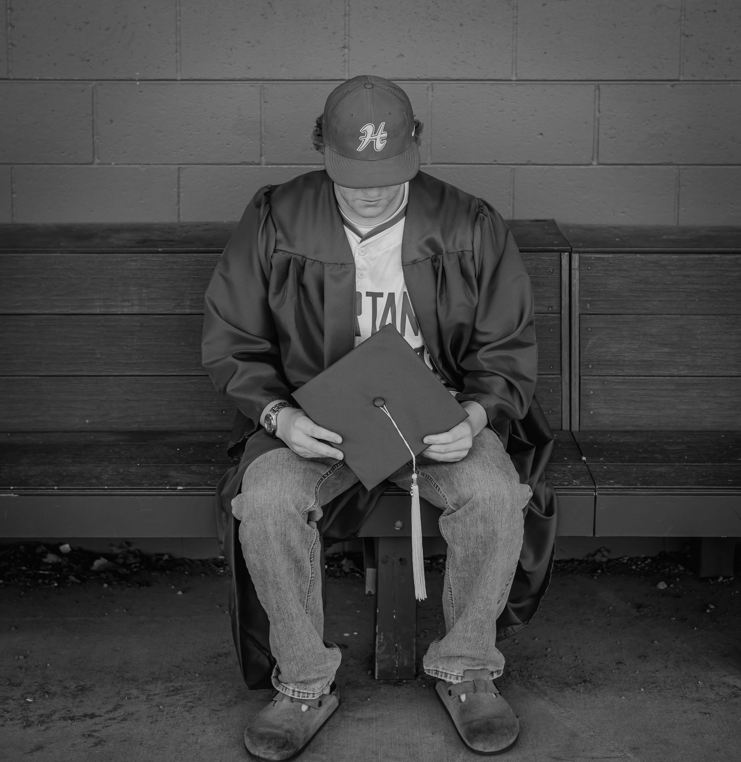Black and white photo of a graduate sitting alone on a bench, head bowed, holding a cap and diploma.