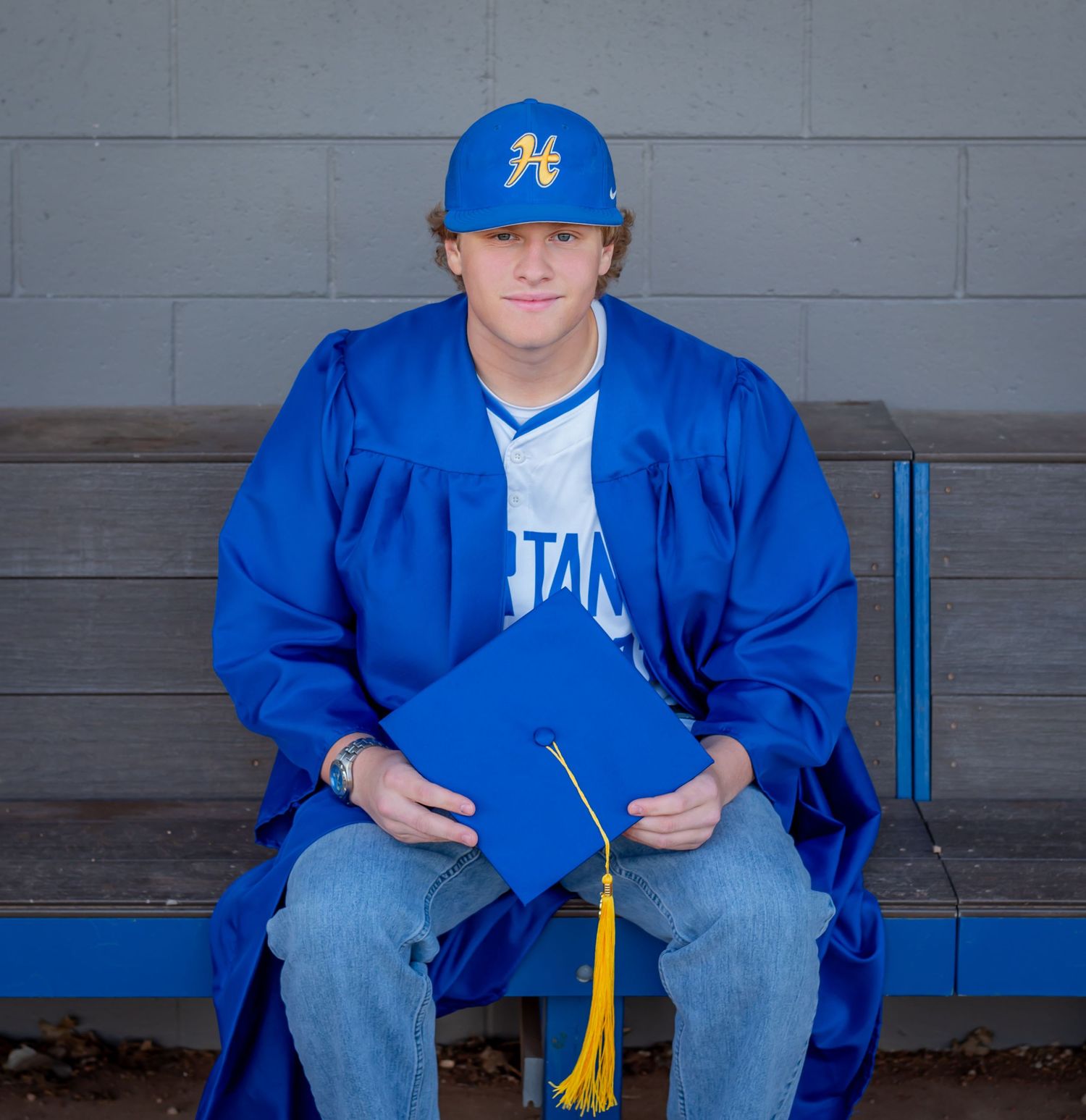 Young man in blue graduation gown and cap holding mortarboard with yellow tassel, seated on bench.