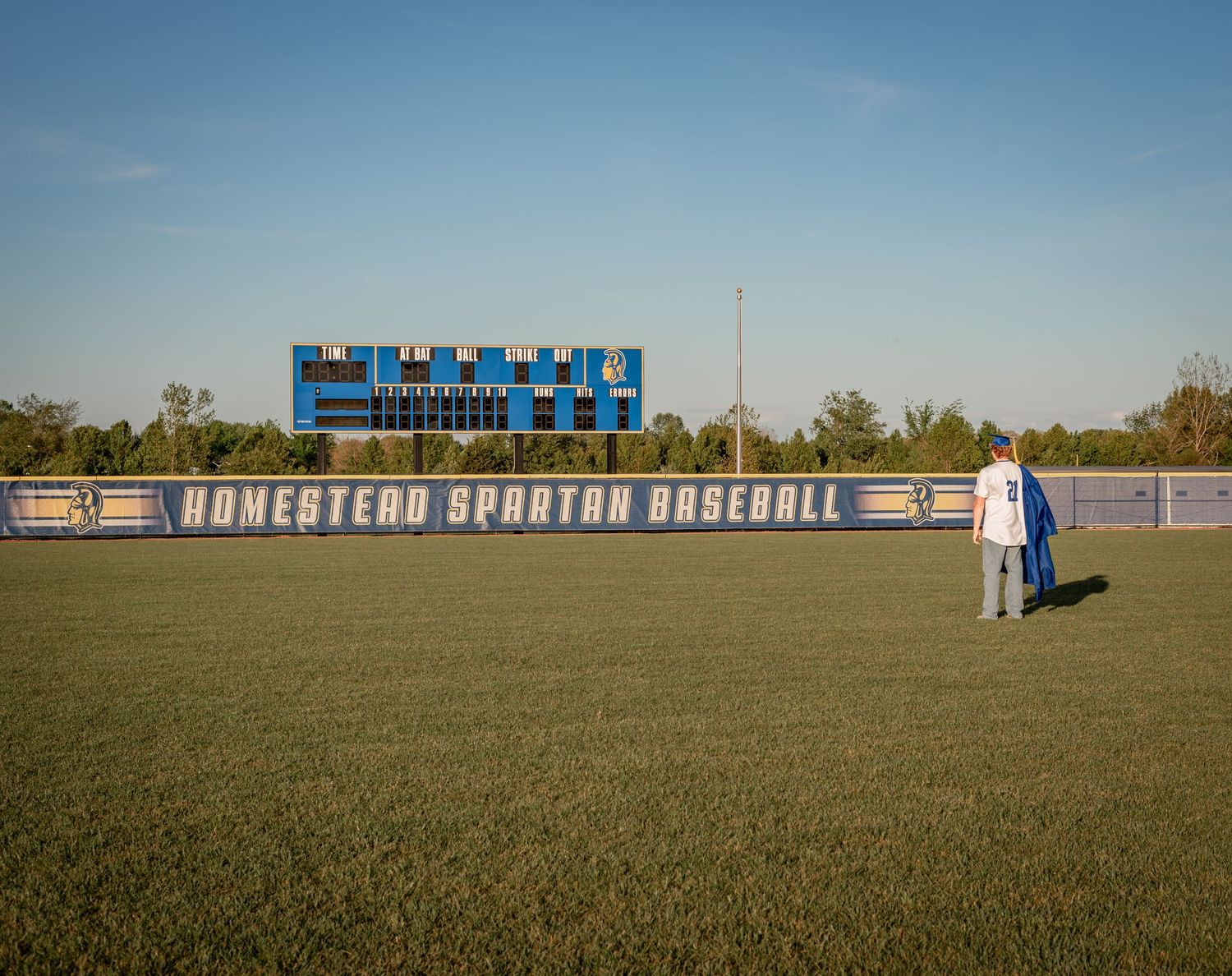 Two people stand on the green field of Homestead Spartan Baseball stadium under a clear blue sky.