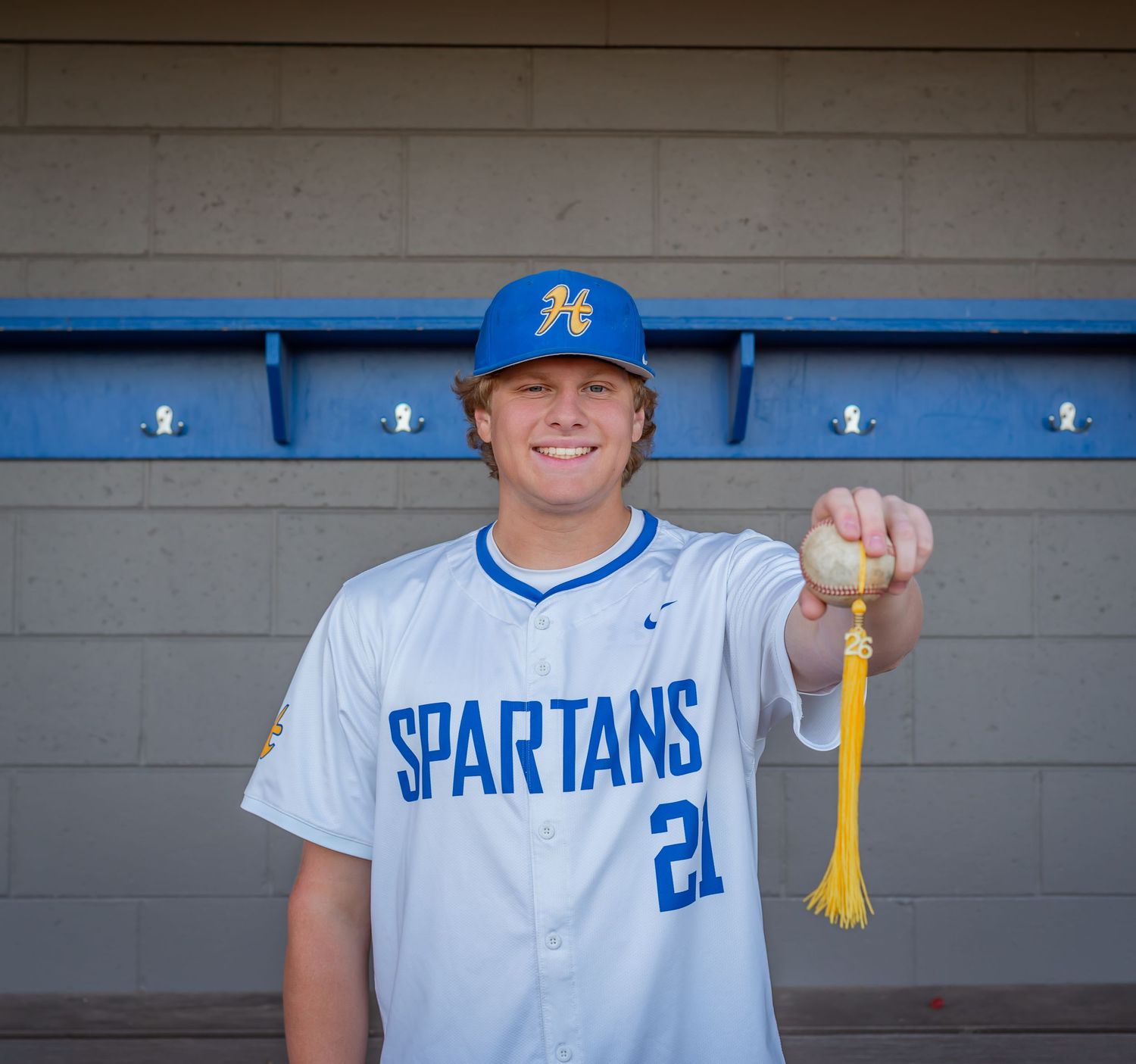Young baseball player in Spartans jersey #21 holding a ball with tassel in dugout wearing blue cap.