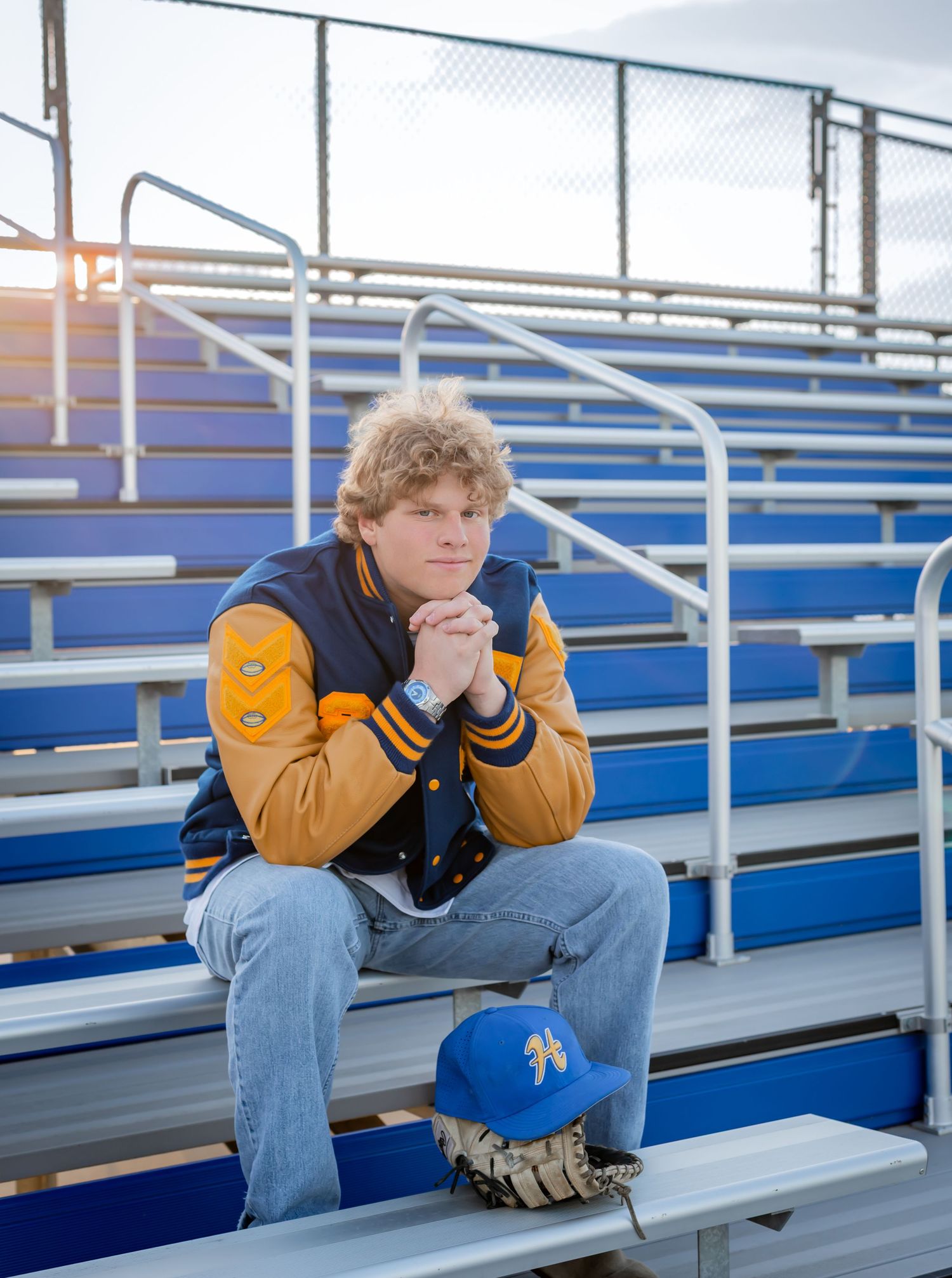 Teen boy in letterman jacket sits on blue bleachers with baseball glove and cap at a sports field.