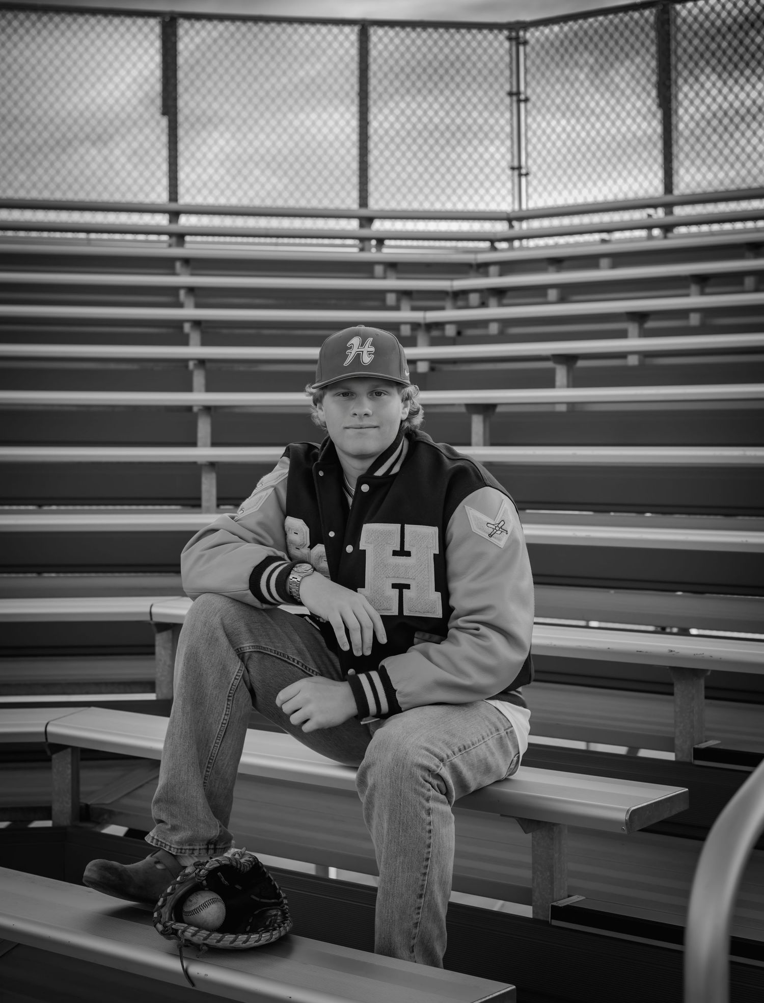 Young man in varsity jacket and baseball cap sits on bleachers holding a glove, black and white photo.