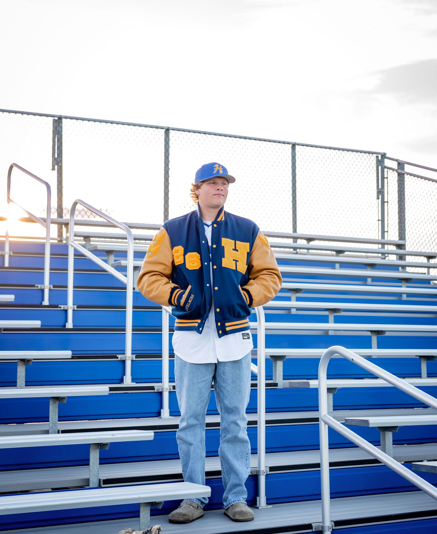 Young man in blue and gold varsity jacket and cap standing on blue bleachers at a sports field.
