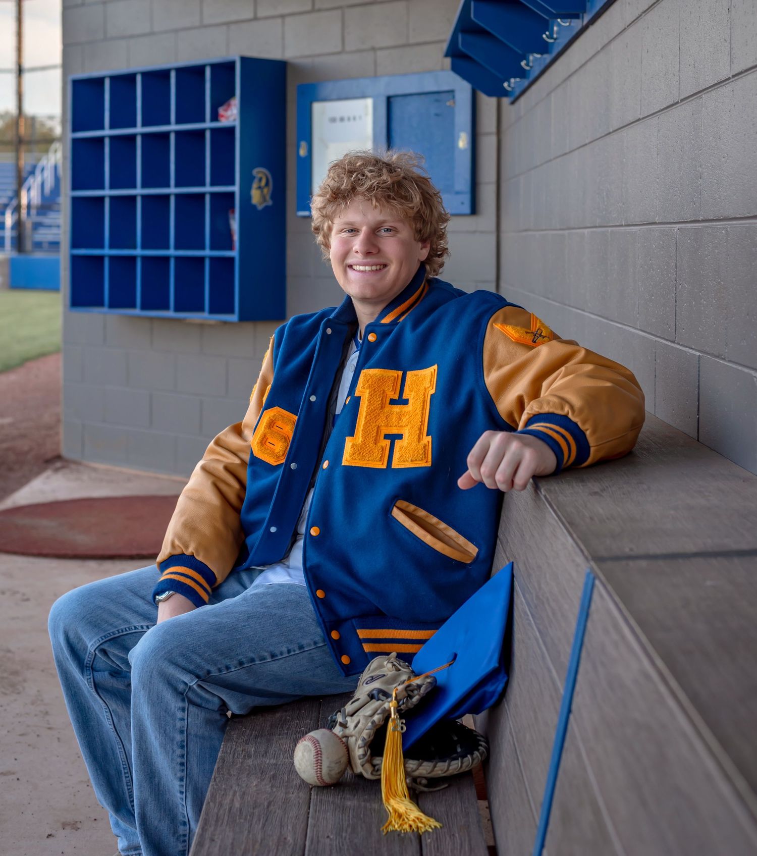 Young man in blue and gold varsity jacket sitting in a baseball dugout with a glove and ball.