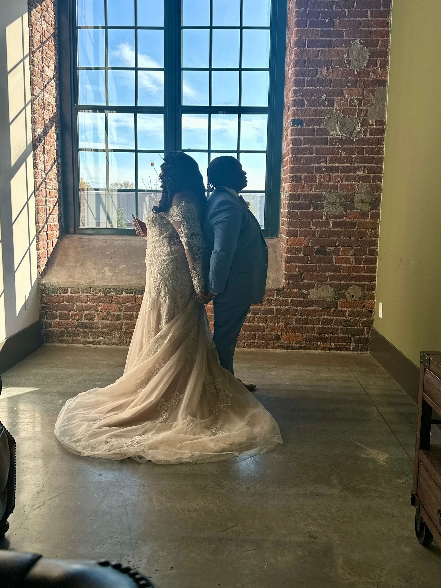 A bride in a flowing lace gown and companion stand silhouetted against large industrial windows in a brick-walled room.