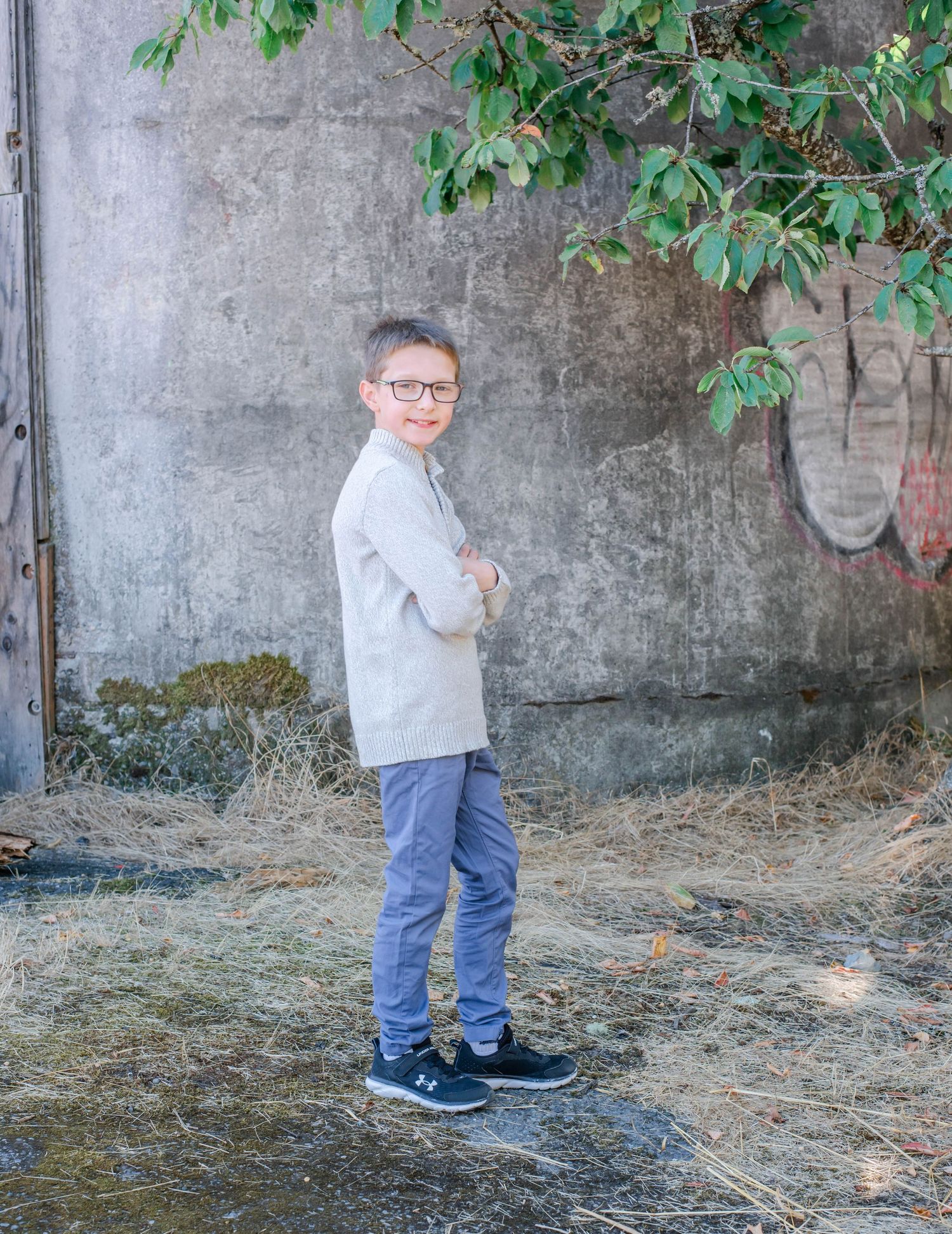 Young boy in gray sweater and jeans plays outdoors near concrete wall and green foliage.