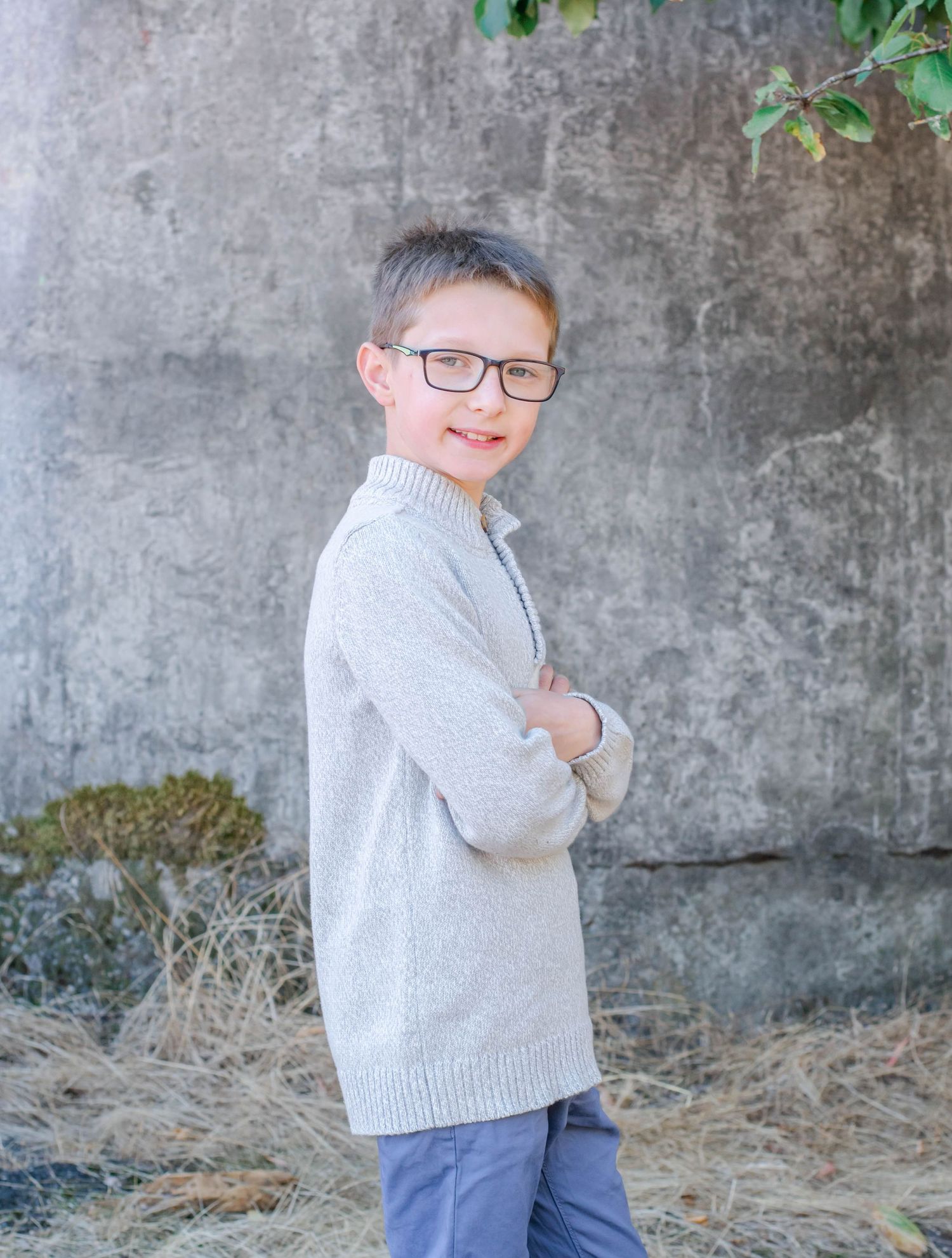 Child in glasses and gray knit sweater poses casually against textured wall background.