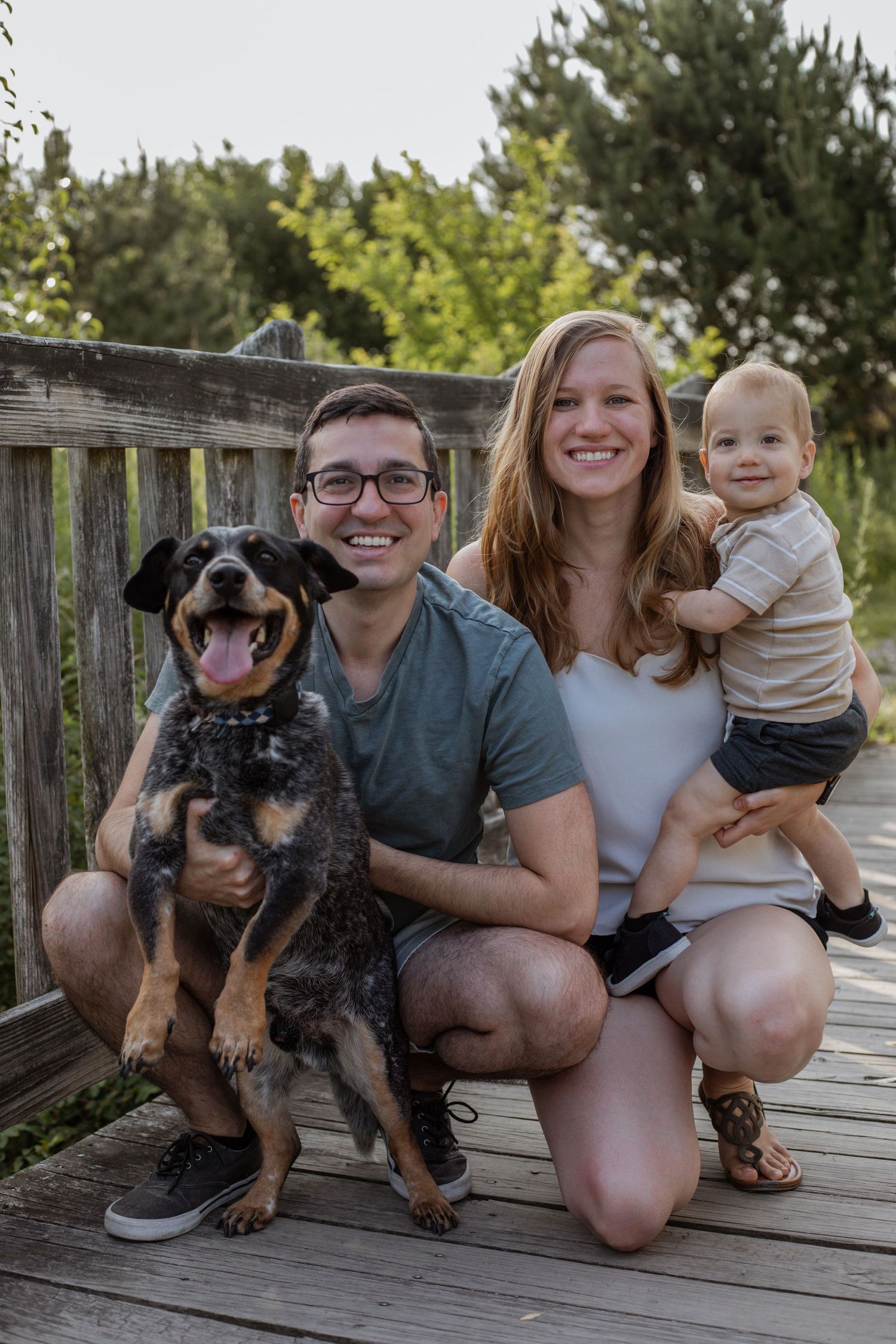 Family poses with their small black and brown puppy on a wooden deck outdoors.