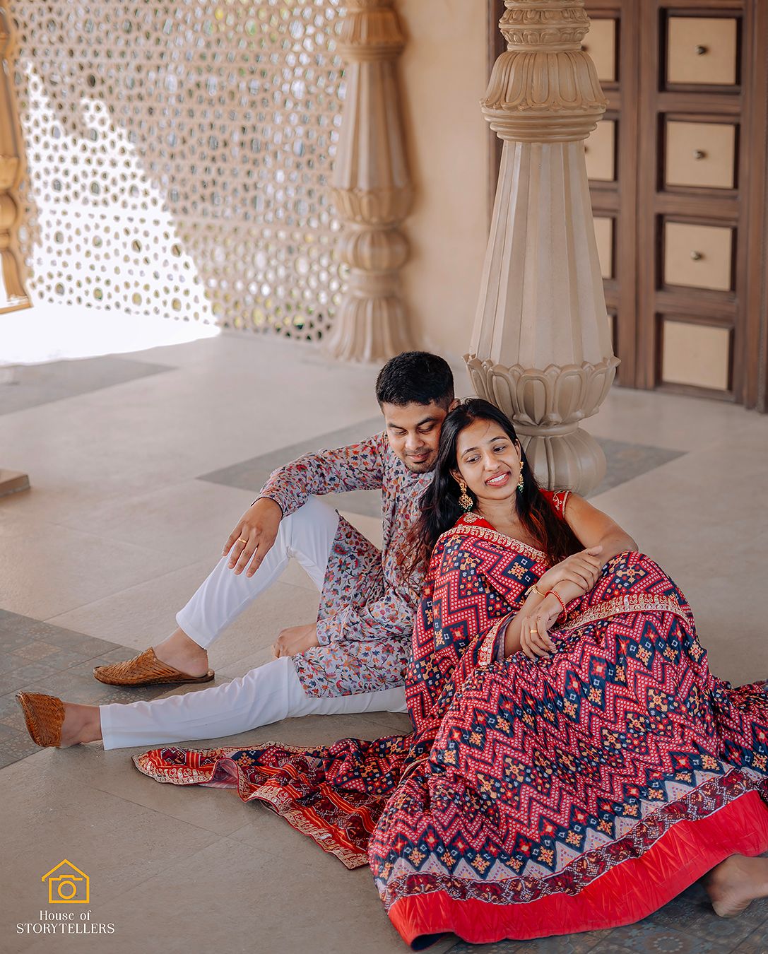 Couple in traditional Indian attire relaxing on ornate floor with decorative pillars.