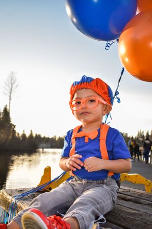 Child in orange beanie and overalls holding blue and orange balloons by a lake at sunset.
