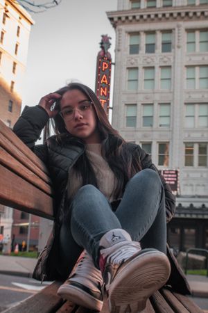 Urban portrait of someone in casual wear sitting on a bench near a vintage theater sign.