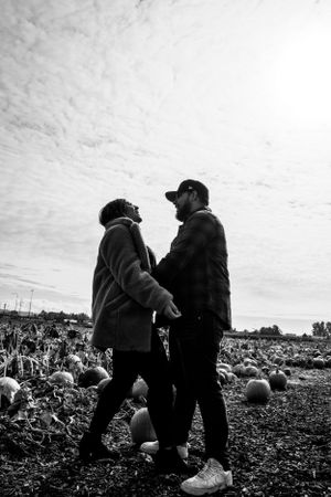 Black and white photo of couple standing in pumpkin field under cloudy sky.