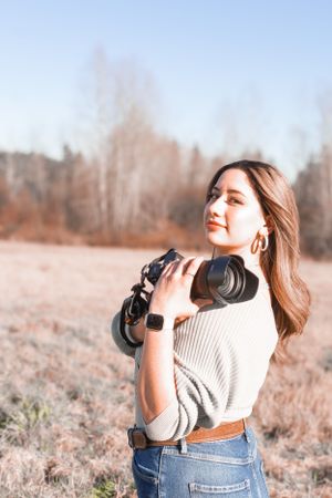Professional photographer in cozy sweater holding camera gear outdoors in autumn field.