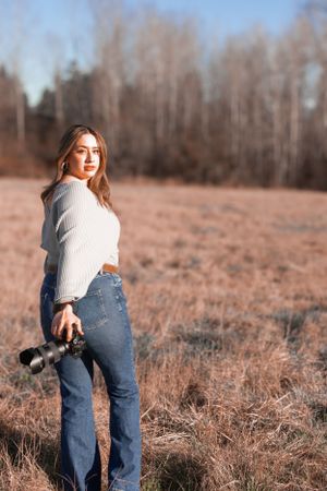 Photographer in white sweater and jeans poses with camera equipment in golden field.