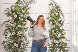 Photographer with camera posing between floral arches in bright indoor setting.