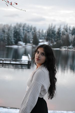 Winter portrait of someone in white sweater posing by a snow-covered lake with evergreen trees.