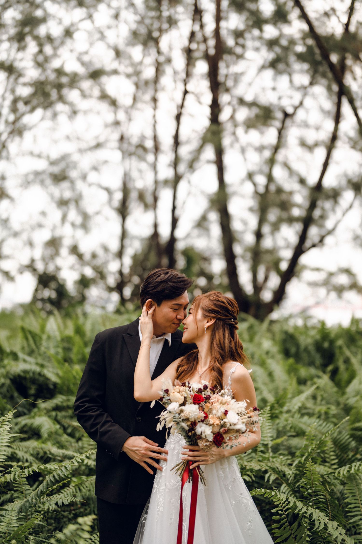 Bride in white lace gown holds bouquet with red ribbons as couple embraces in forest.