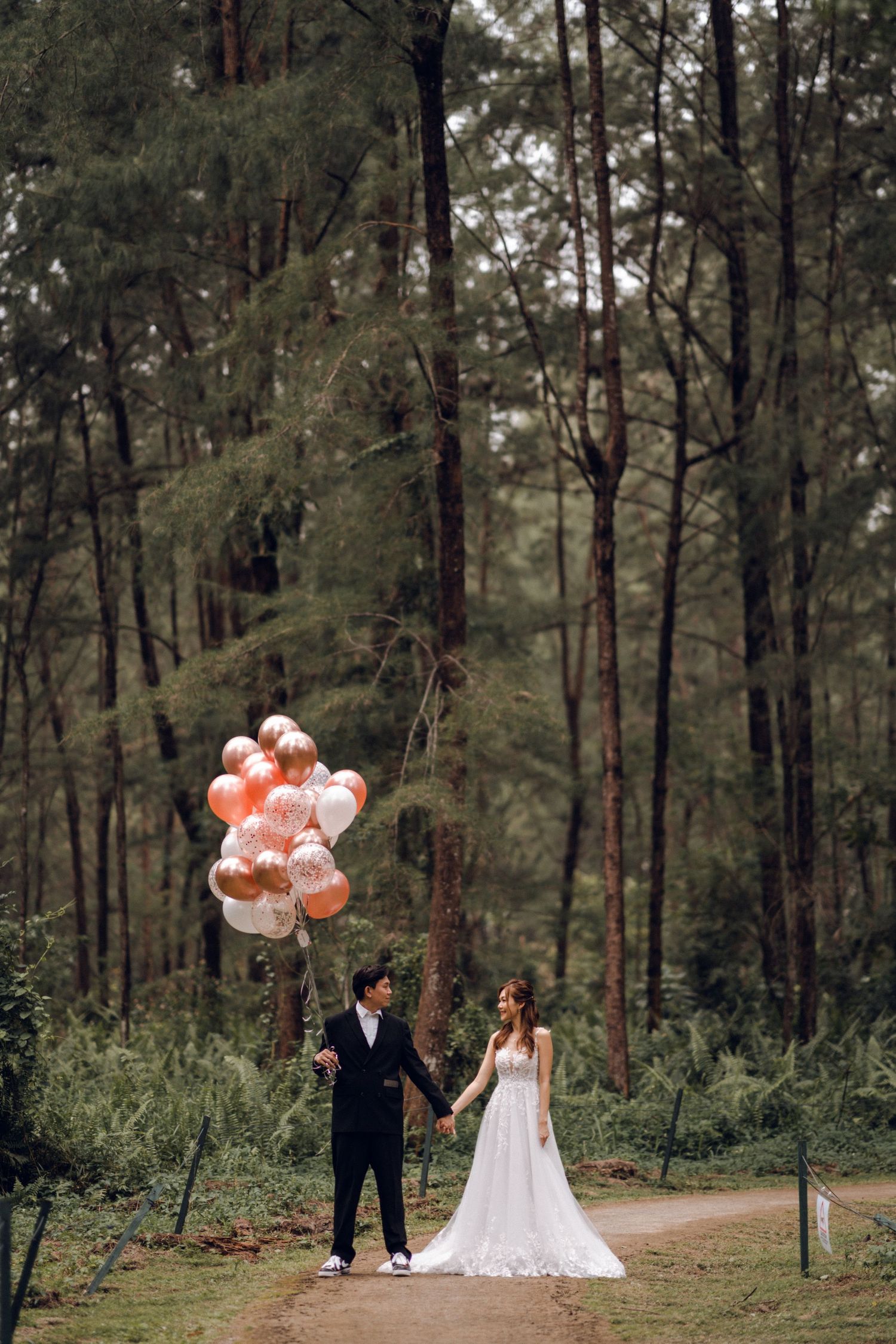 Wedding couple holds peach and white balloons in serene forest setting.