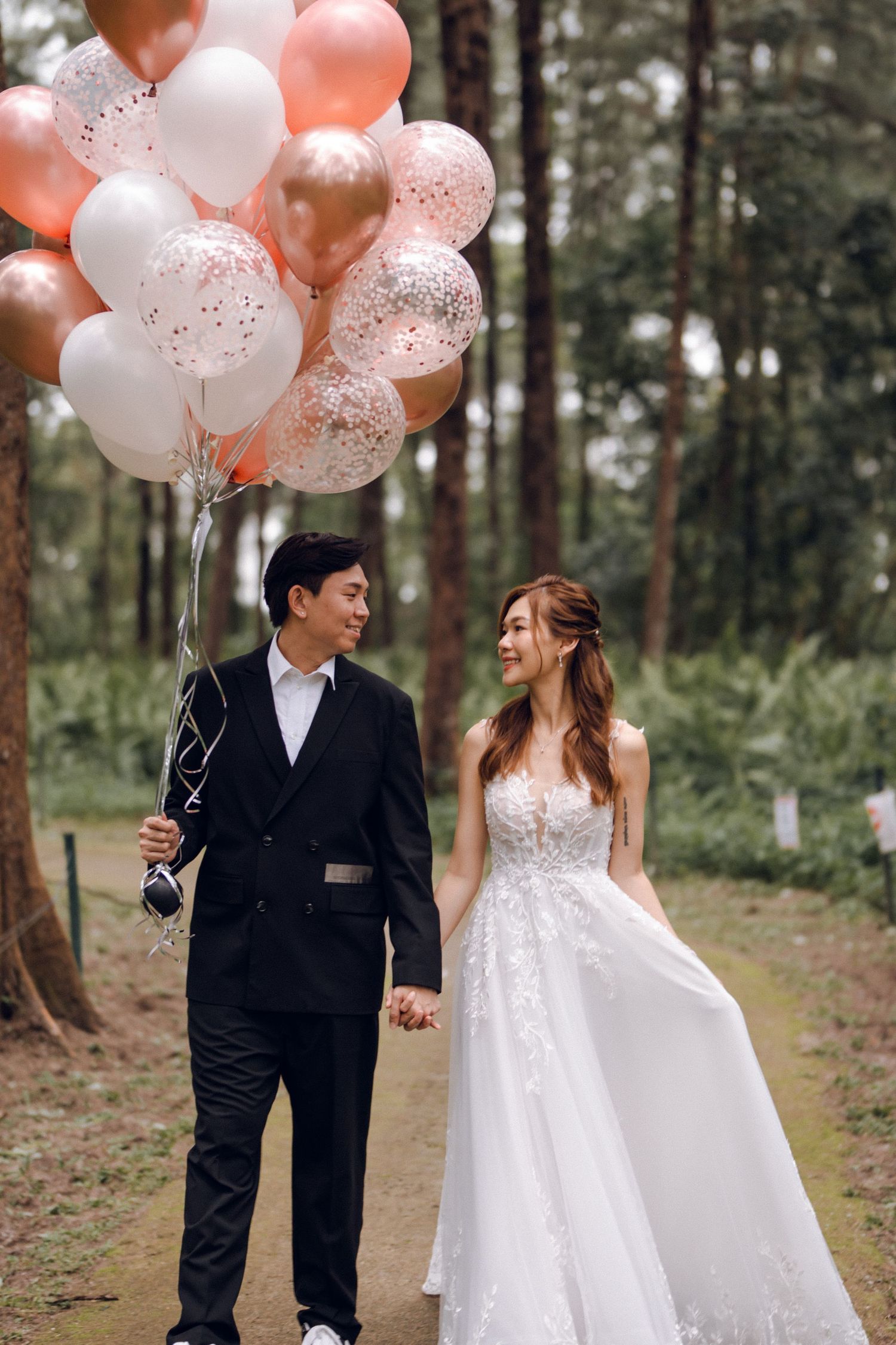 Wedding couple poses with peach and white balloons in a serene forest setting.