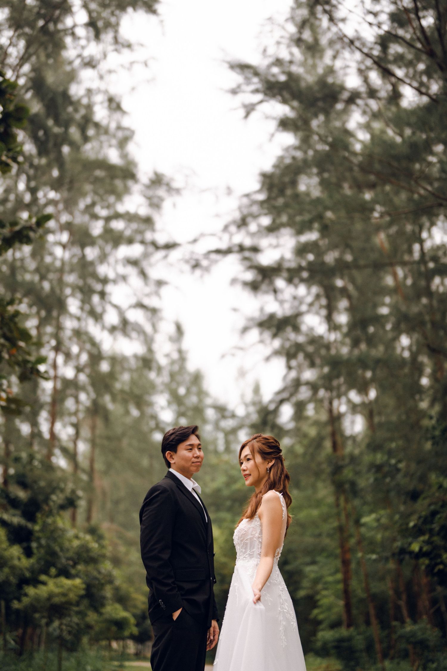 A romantic wedding couple shares a tender moment on a tree-lined path in a forest setting.