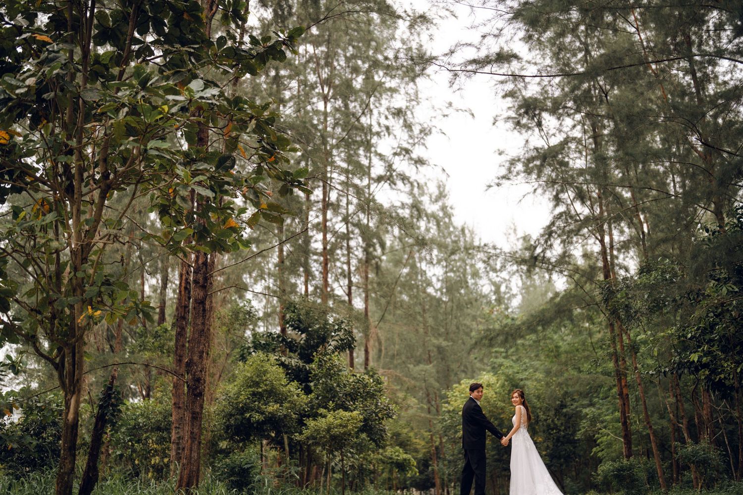 A wedding couple shares a joyful moment surrounded by tall evergreen trees.