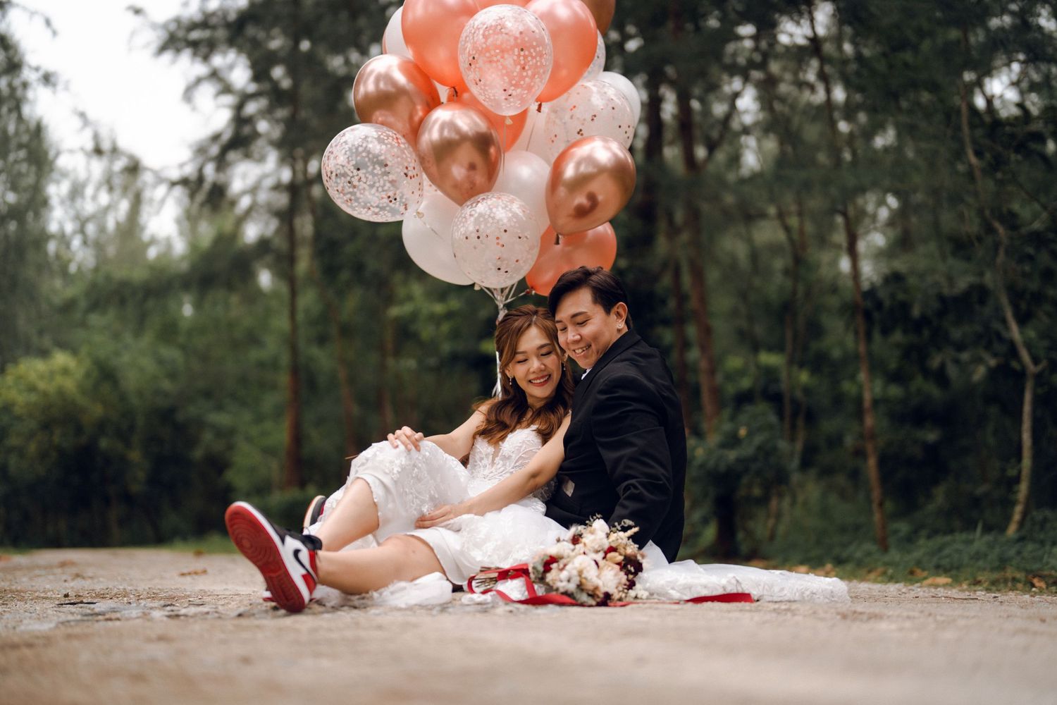 Newlyweds pose playfully with peach and white balloons on a forest path.