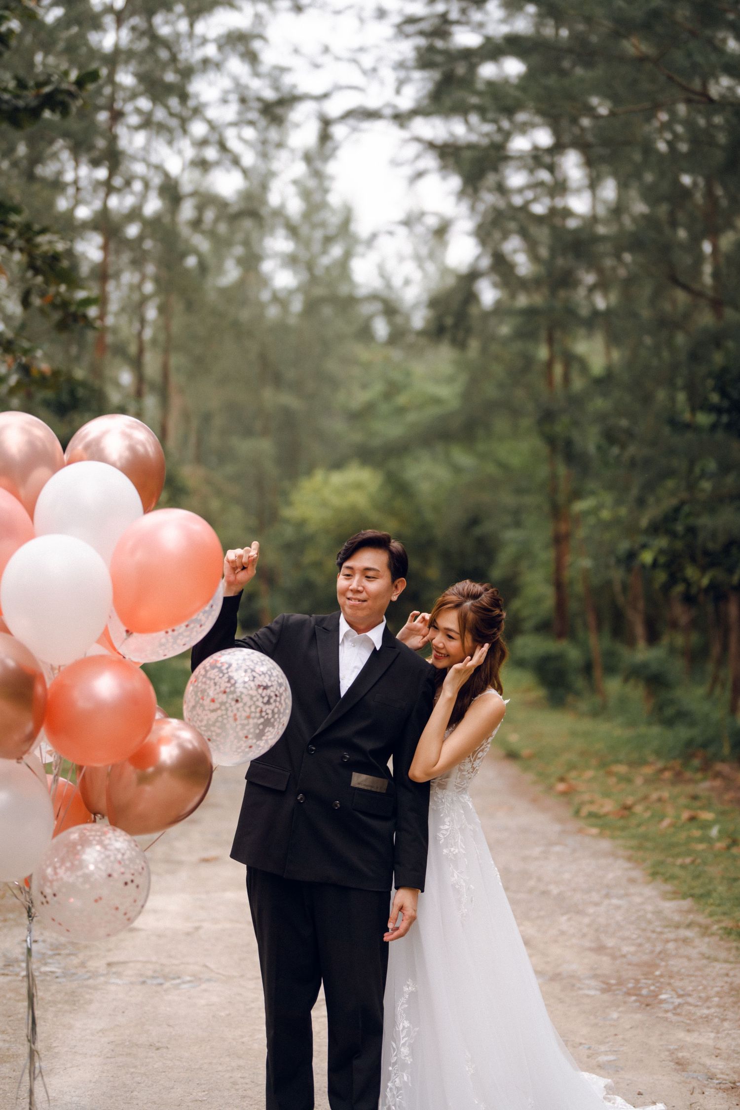 A couple celebrates with peach and white balloons along a wooded trail.