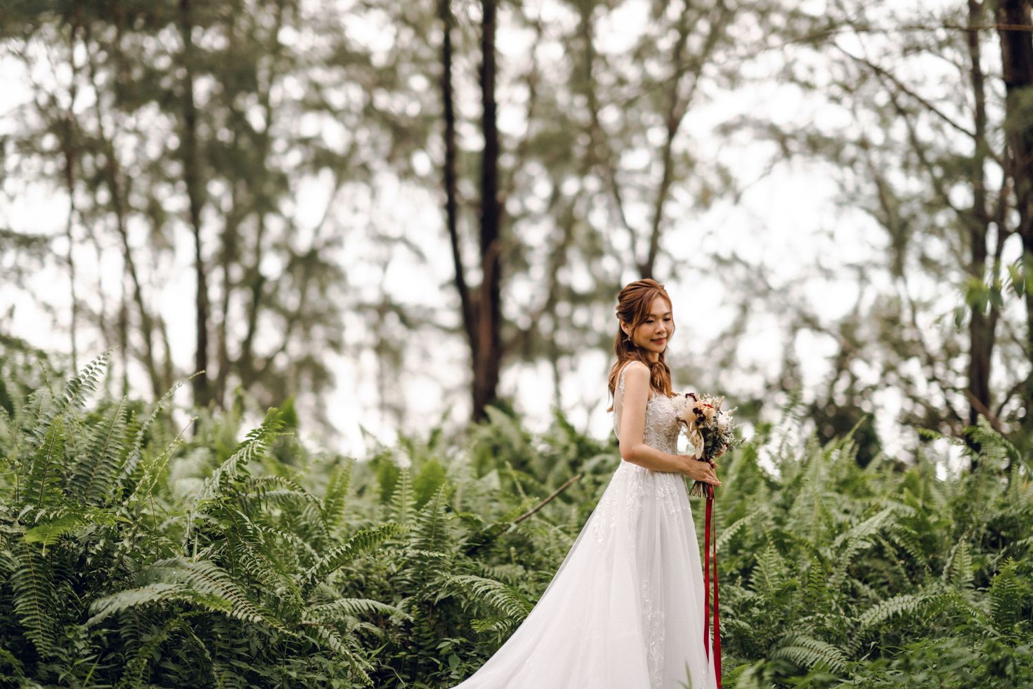 Bride in flowing white dress poses elegantly among lush forest ferns.