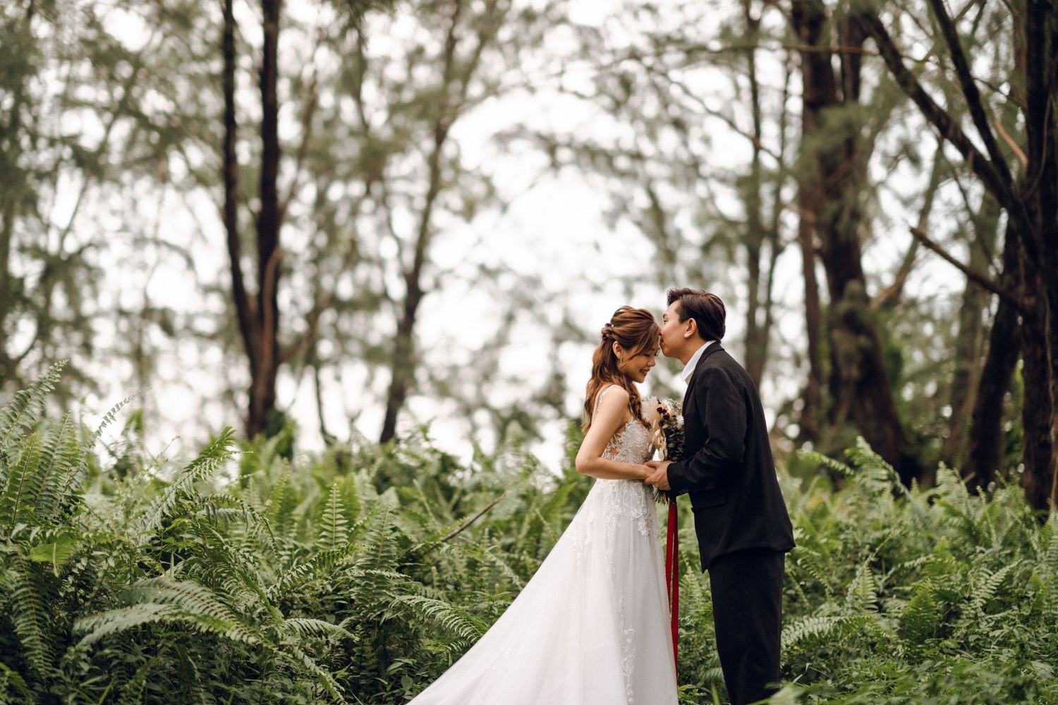 Romantic wedding couple poses in a lush forest setting with tall trees and ferns.