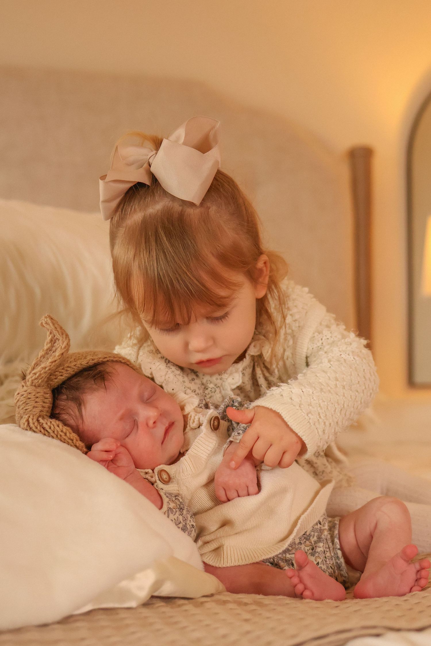 A tender moment captured as big sister in white dress gently touches newborn sibling on soft bedding in warm light.