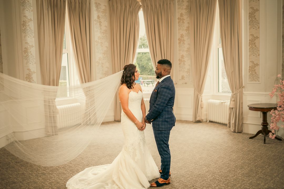 Romantic full-length portrait of newlyweds in elegant room with flowing curtains.