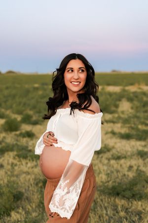 A maternity photo series showing a pregnant woman in a white lace dress standing in a grassy field at sunset.