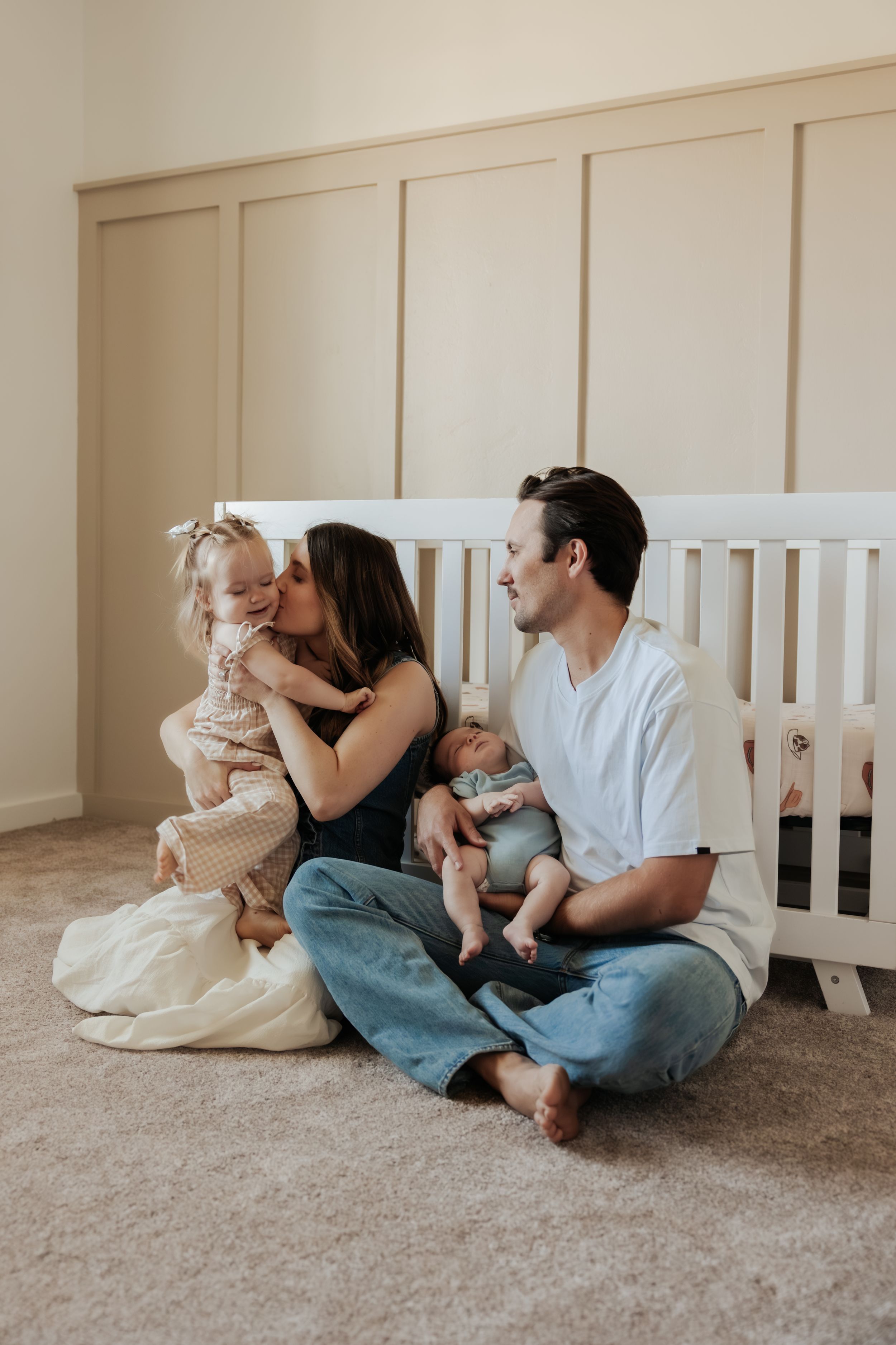 Family lying together on carpeted floor near white cribs in casual home setting showing tender connection.
