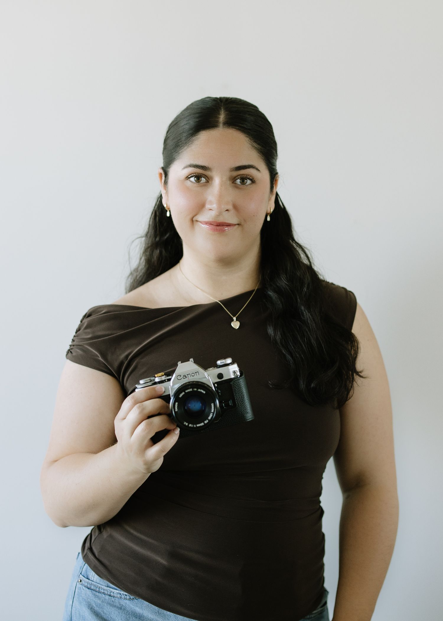A Northern California photographer in a brown t-shirt poses with a professional DSLR camera against a light gray wall.