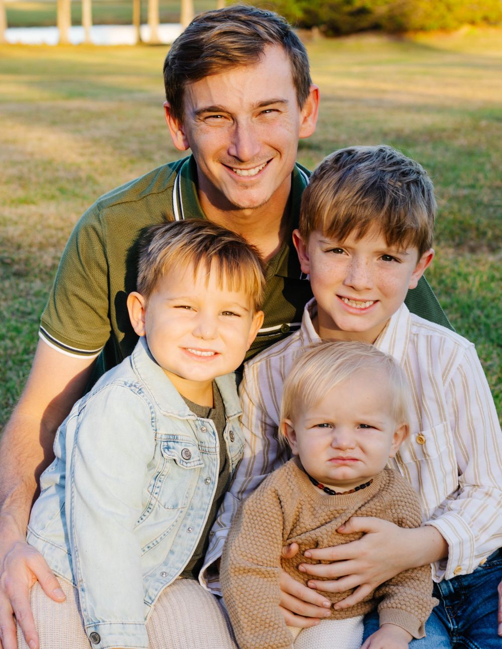 Family portrait outdoors in warm sunset light with four people wearing casual clothing against a grassy background.