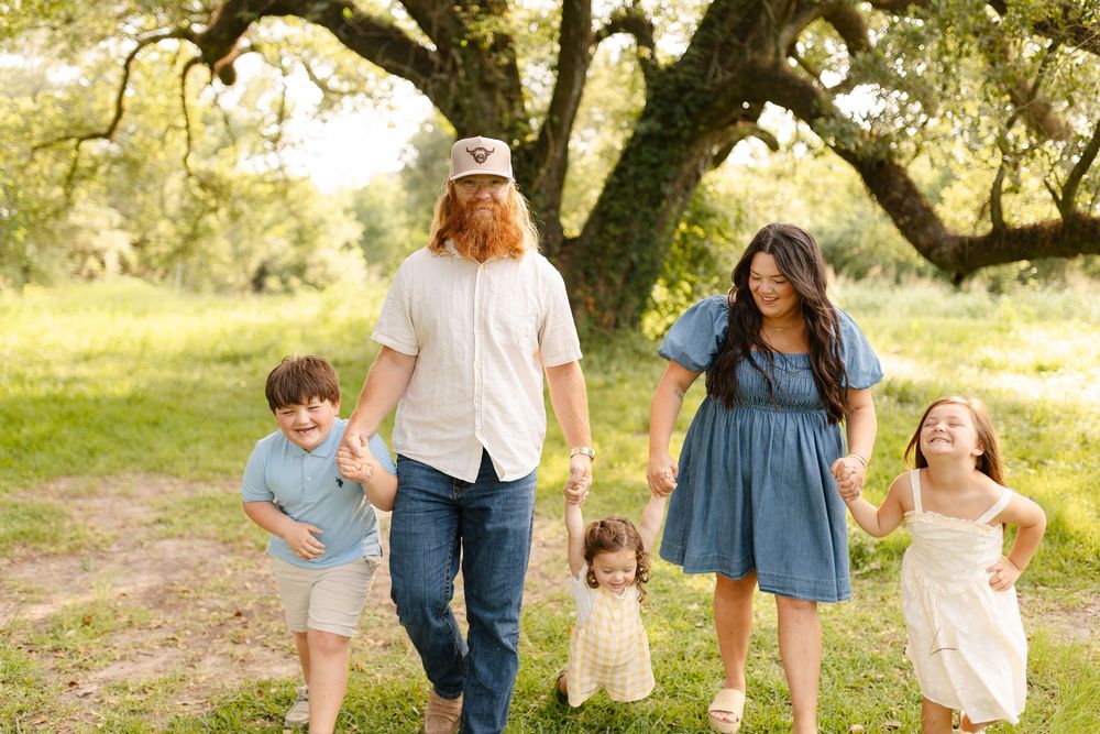 Happy family of five walking hand-in-hand outdoors on green grass beneath a large oak tree on a sunny day.