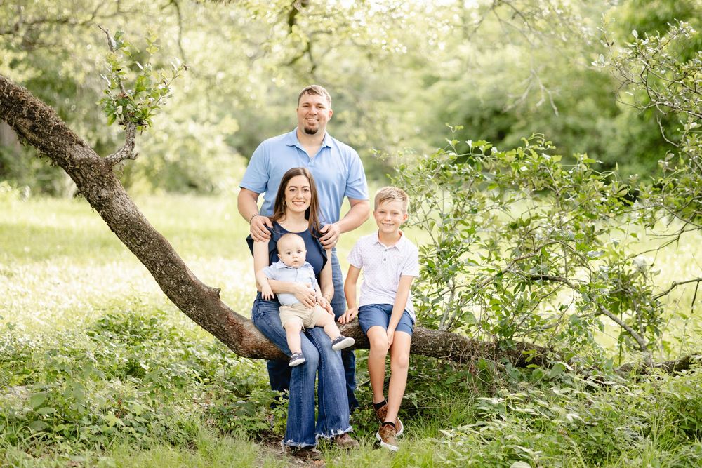 Family of four poses outdoors among blooming trees, parents with two children sitting on a fallen branch in a lush green setting.