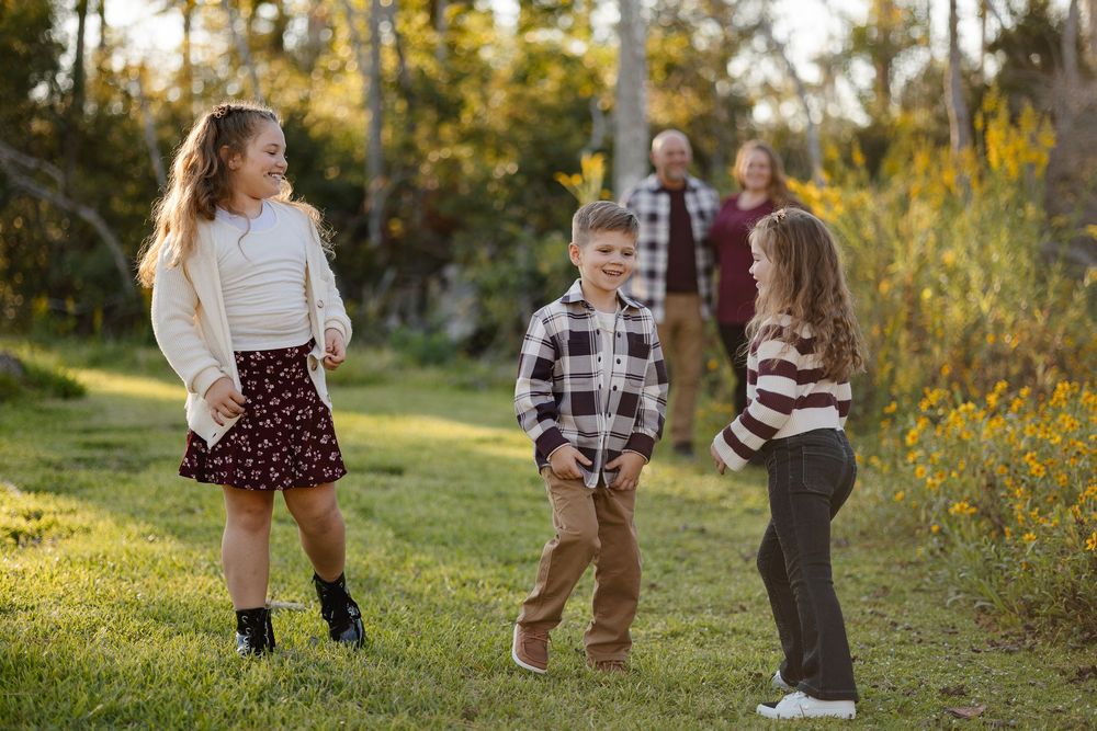 Family with three children playing outdoors in a sunny park surrounded by trees and yellow wildflowers.