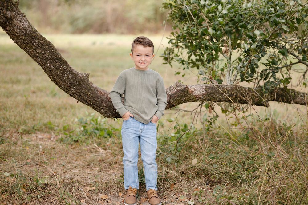 Young boy in green sweater and jeans poses by a leaning tree branch in an outdoor field setting.