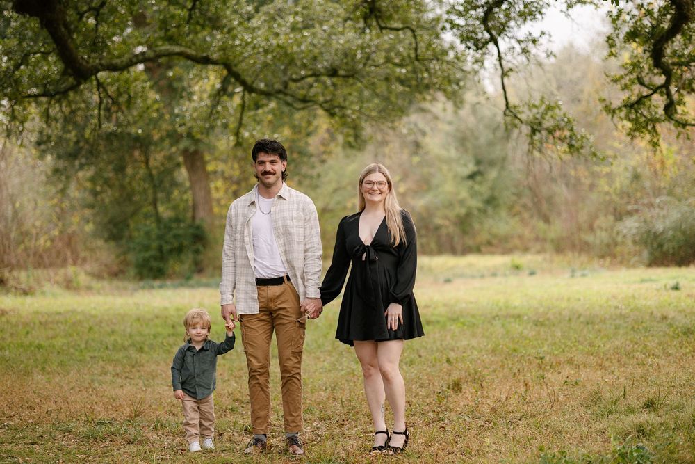 Family of three posing outdoors in a lush green field, parents holding hands with toddler, surrounded by trees.